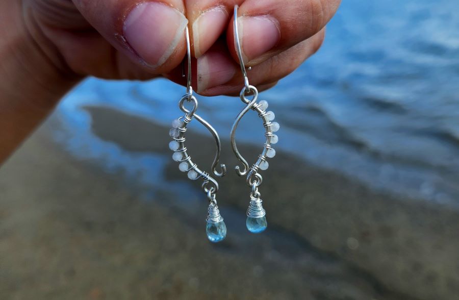 Beach-inspired earrings with aquamarine drops and wire-wrapped moonstone in front of ocean water