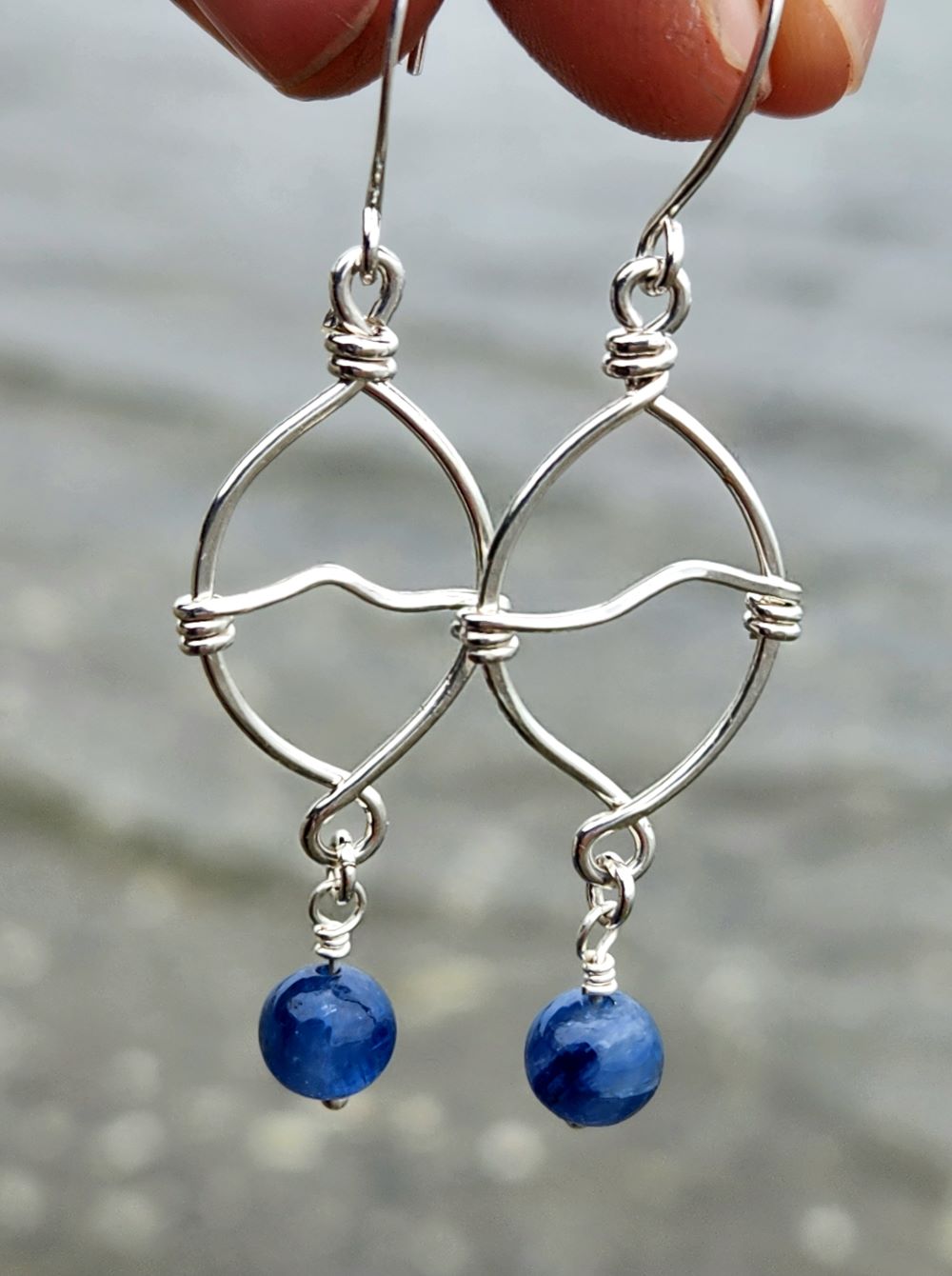 Handmade sterling silver wave earrings with natural kyanite gemstone in front of ocean waves